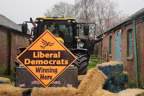Ed Davey waves from tractor with Lib Dem sign, having smashed blue wall of bales